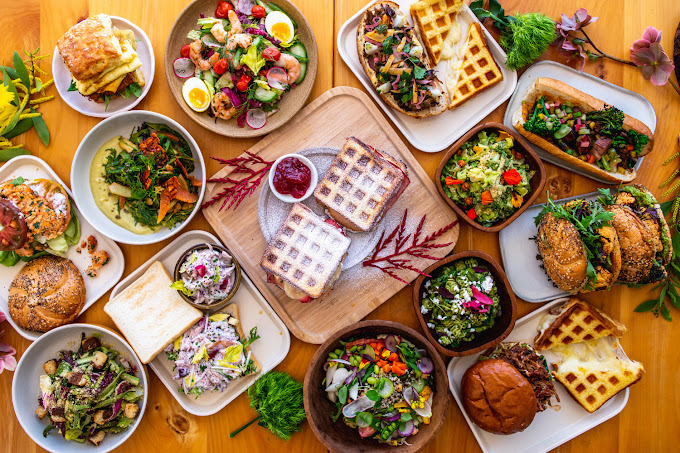 Top view of a layout of numerous dishes on a wooden table. It features waffles, salads, and sandwiches.