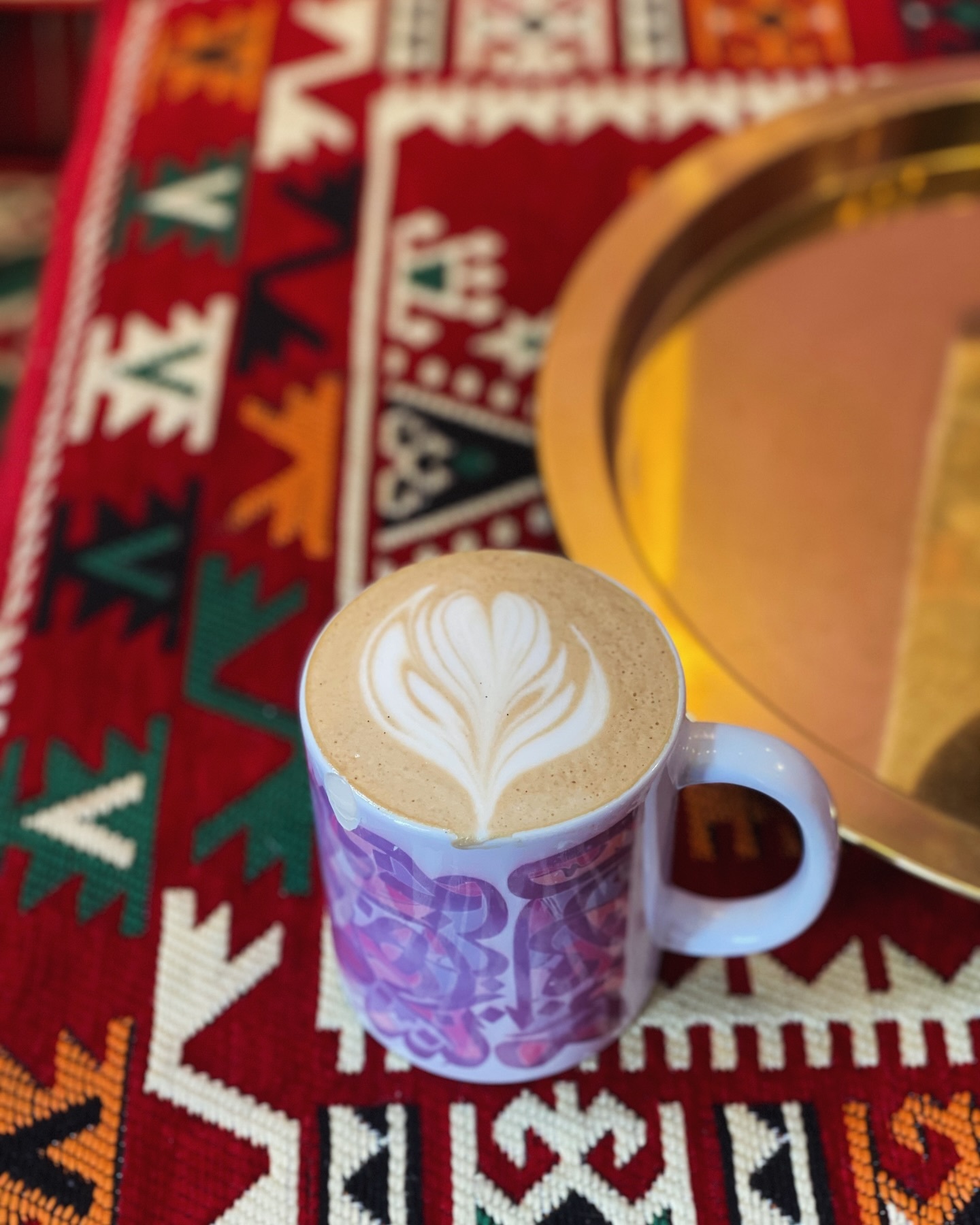 Hot latte with heart foam art in a blue and purple mug. The background is red, green, white, and orange woven piece. A shiny gold plate sits off to the right edge of the image.