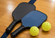 Two pickleball paddles resting on a hardwood gym floor beside two bright yellow pickleballs.