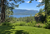 Lush green lawn overlooking a forest, blue lake, and distant mountains beneath a clear sunny sky with no people visible.