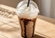 Frozen chocolate coffee drink in a clear plastic cup with chocolate drizzle, whipped cream, and a black straw on a wooden tabletop.