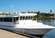 White passenger boat docked along a calm waterfront under a bright blue sky with gently rippling water and a softly blurred shoreline in the background.