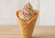 Chocolate soft serve in a waffle cone topped with colorful rainbow sprinkles against a clean, softly blurred neutral background.