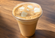 Creamy iced coffee in a clear plastic cup with a flat lid and ice cubes, photographed on a warm wooden table in natural light.