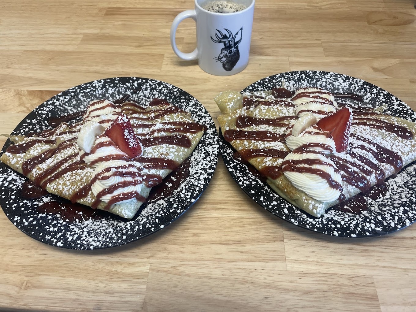 A view of two black plates both serving up a crepe with whipped cream, strawberries and chocolate drizzle.