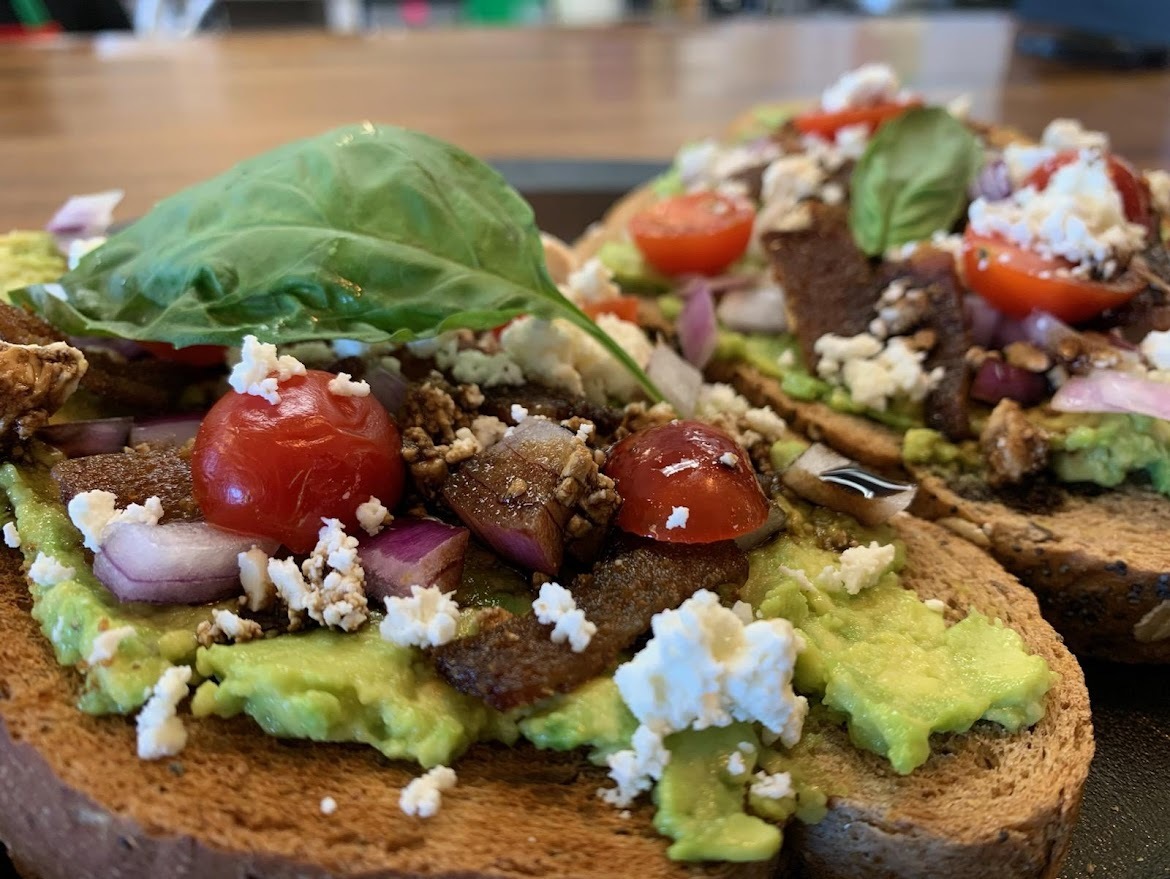 A view of a black plate consisting of two slices of avocado toast topped with onions, tomatoes, feta cheese, and garnish.