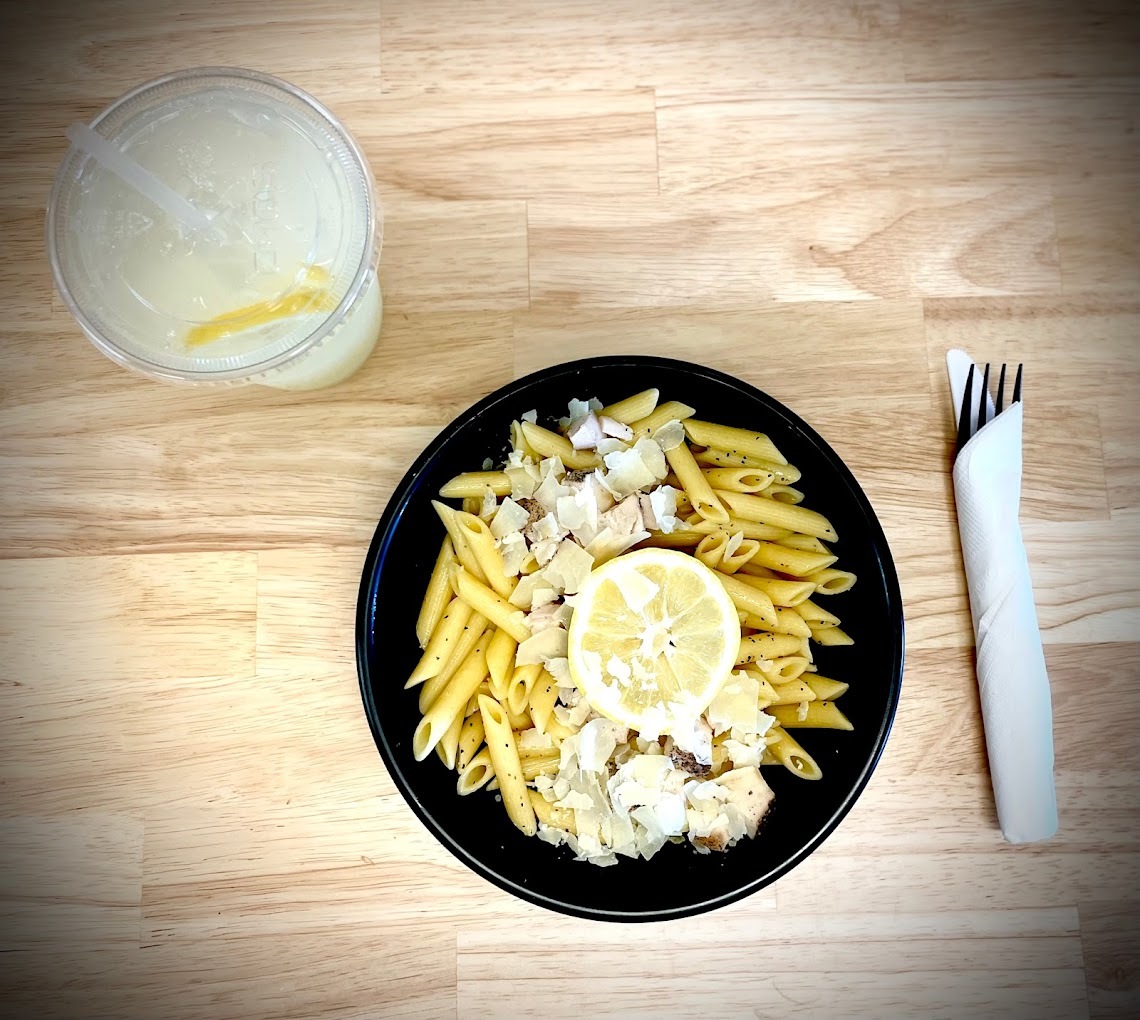 A black plate consisting of pasta topped with a lemon along side a lemonade drink.