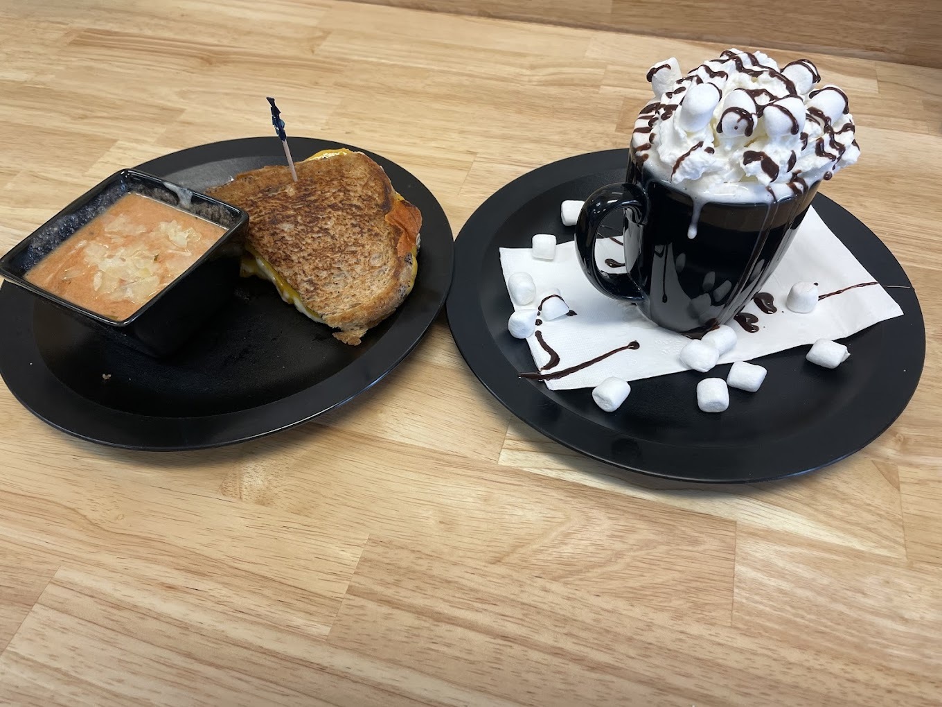 A black plate consisting of a panini sandwich and another small round black plate with a mug topped with whipped cream, marshmallows, and chocolate drizzle.