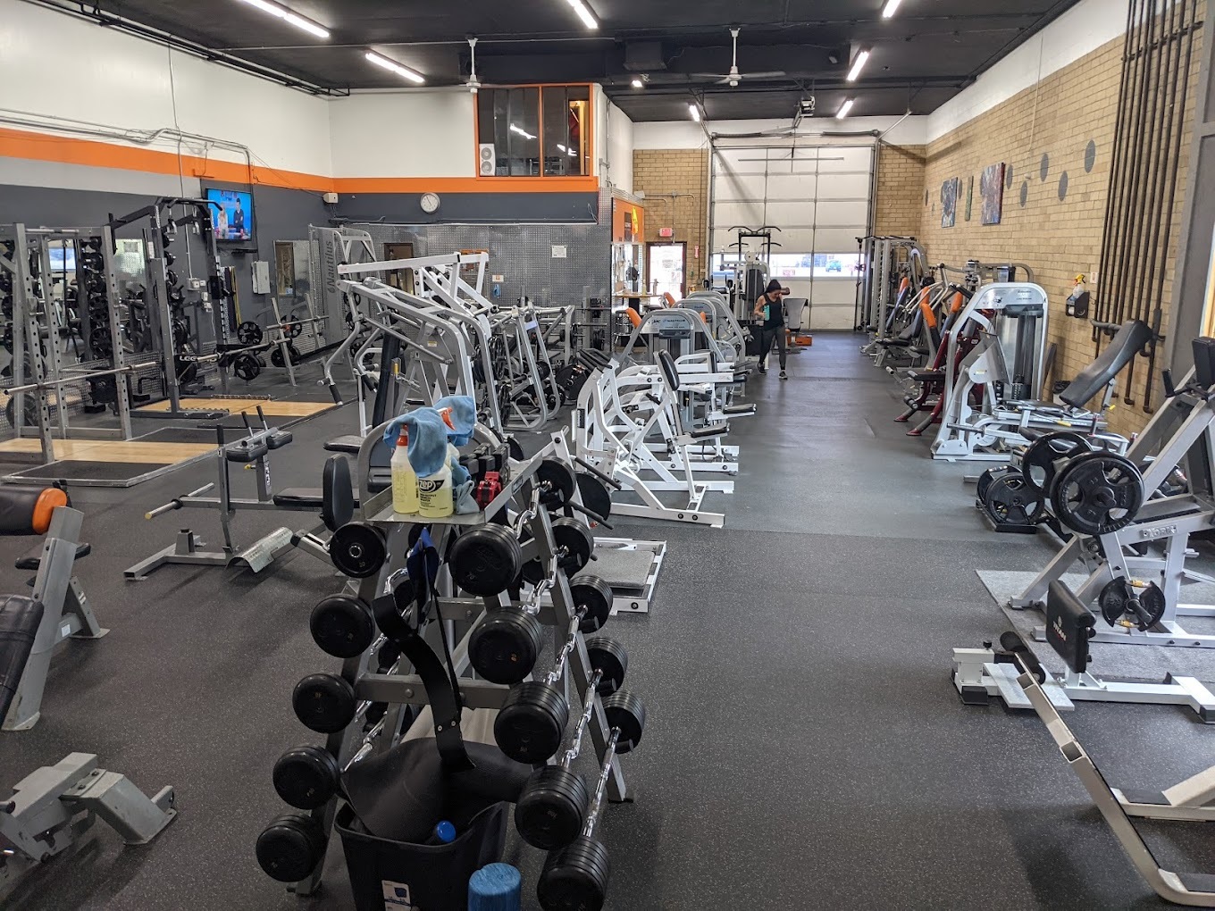 A large brick room with a large garage door on the very back wall, the room is filled with workout equipment, racks of weights, and other cardio and strength equipment.