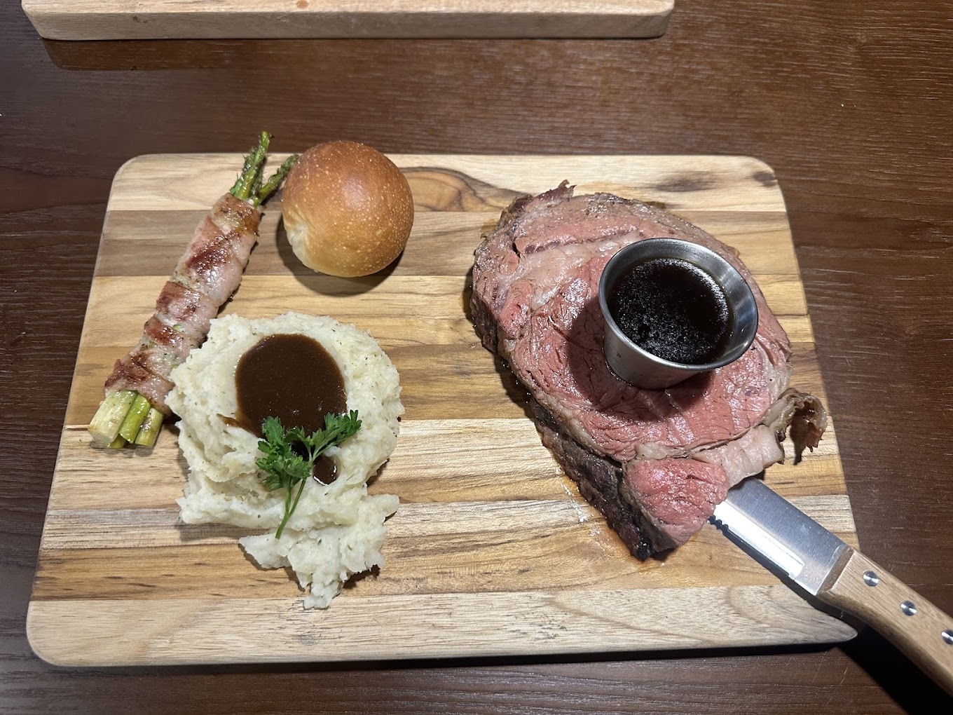 A cutting board consisting of mashed potatoes, and a steak with a metal cup containing a side of sauce, along with a bread roll and asparagus wrapped in bacon.