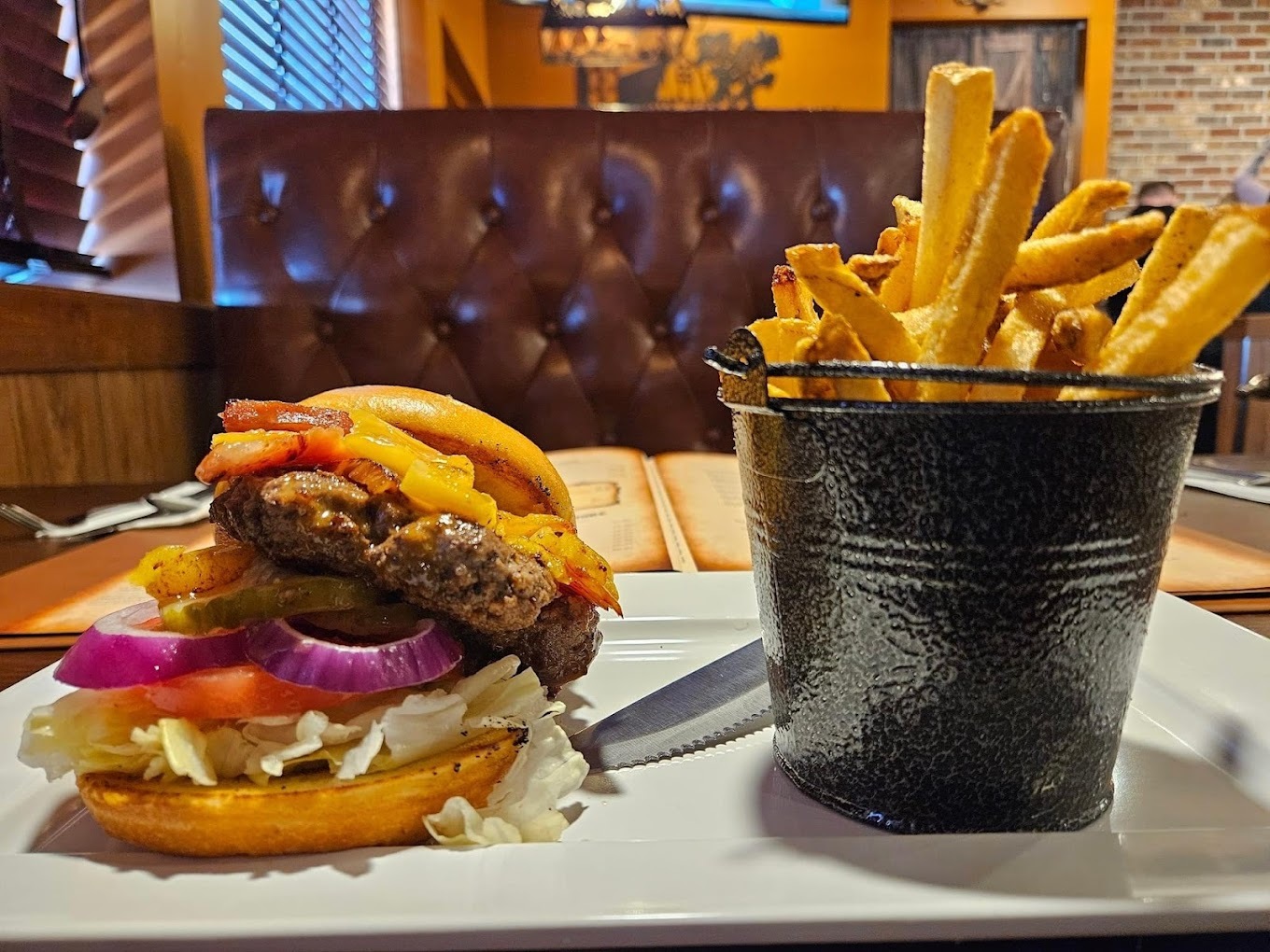 A view of a white plate with a large burger with onion, lettuce, tomato and cheese along with a side of regular cut fries in a black pail with a handle.