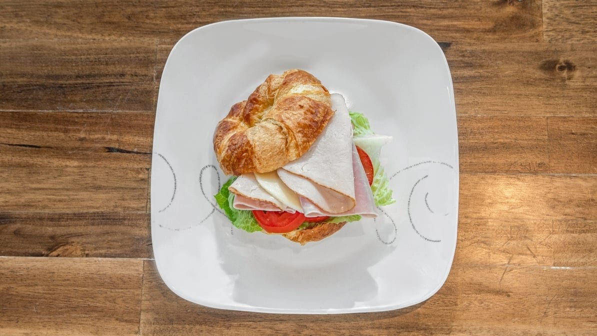 A white plate consisting of a croissant sandwich with meat, lettuce, cheese, and tomato.