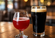Two drinks on a wooden table, including a dark beer in a pint glass and a red fruit beverage in a stemmed glass, with a softly blurred bar background.