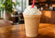 Creamy blended coffee drink topped with whipped cream and a bright red straw in a clear plastic cup on a wooden café table.