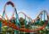 Bright orange roller coaster winding through loops and curves above landscaped greenery and water on a clear day.