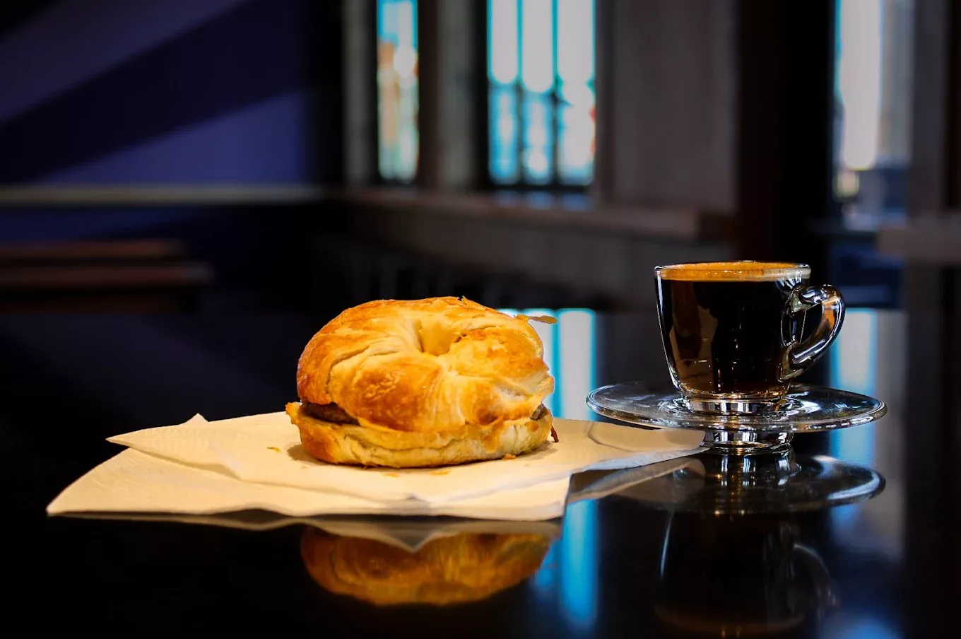 A black table with a white napkin with a croissant sandwich along with a clear coffee mug with an espresso inside.