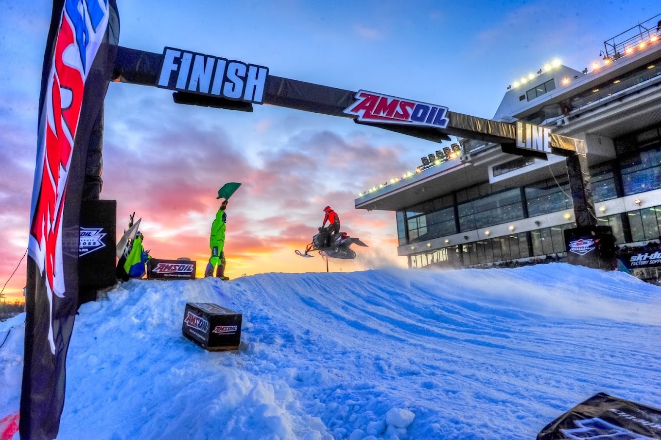 A view of a horse racing track turned into a snowmobiling track with a view of a racer reaching the finish line with a sunset in the background.
