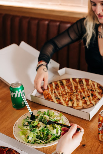 A view of a table with a white box consisting of a pizza and a white plate consisting of a salad, along with two cans of pop off to the side.