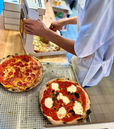A view of two pizzas being prepped by a chef on an conveyer belt along with another pizza being boxed off to the side of the belt.