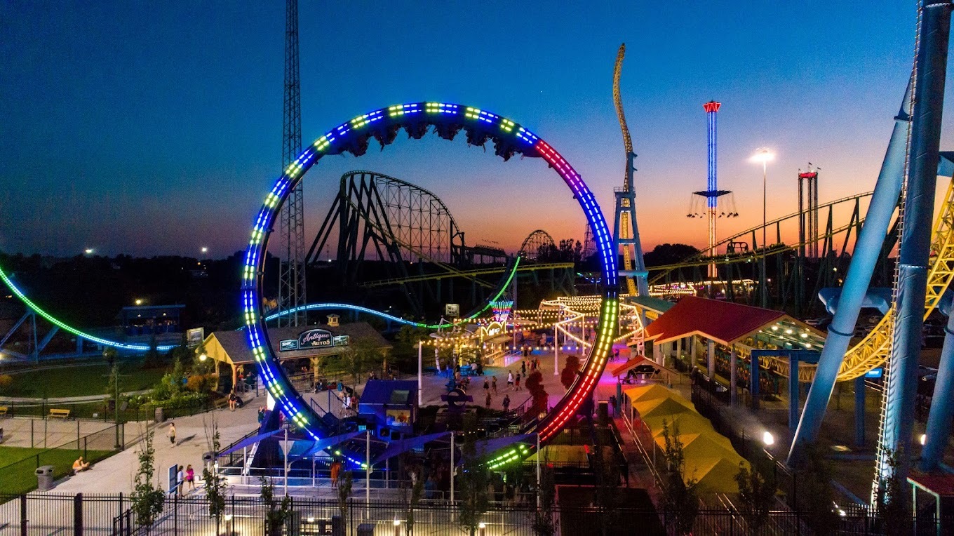 A view of an amusement park at night, colorful lights light up the amusement rides and roller coasters, a blue and orange sunset is in the background.