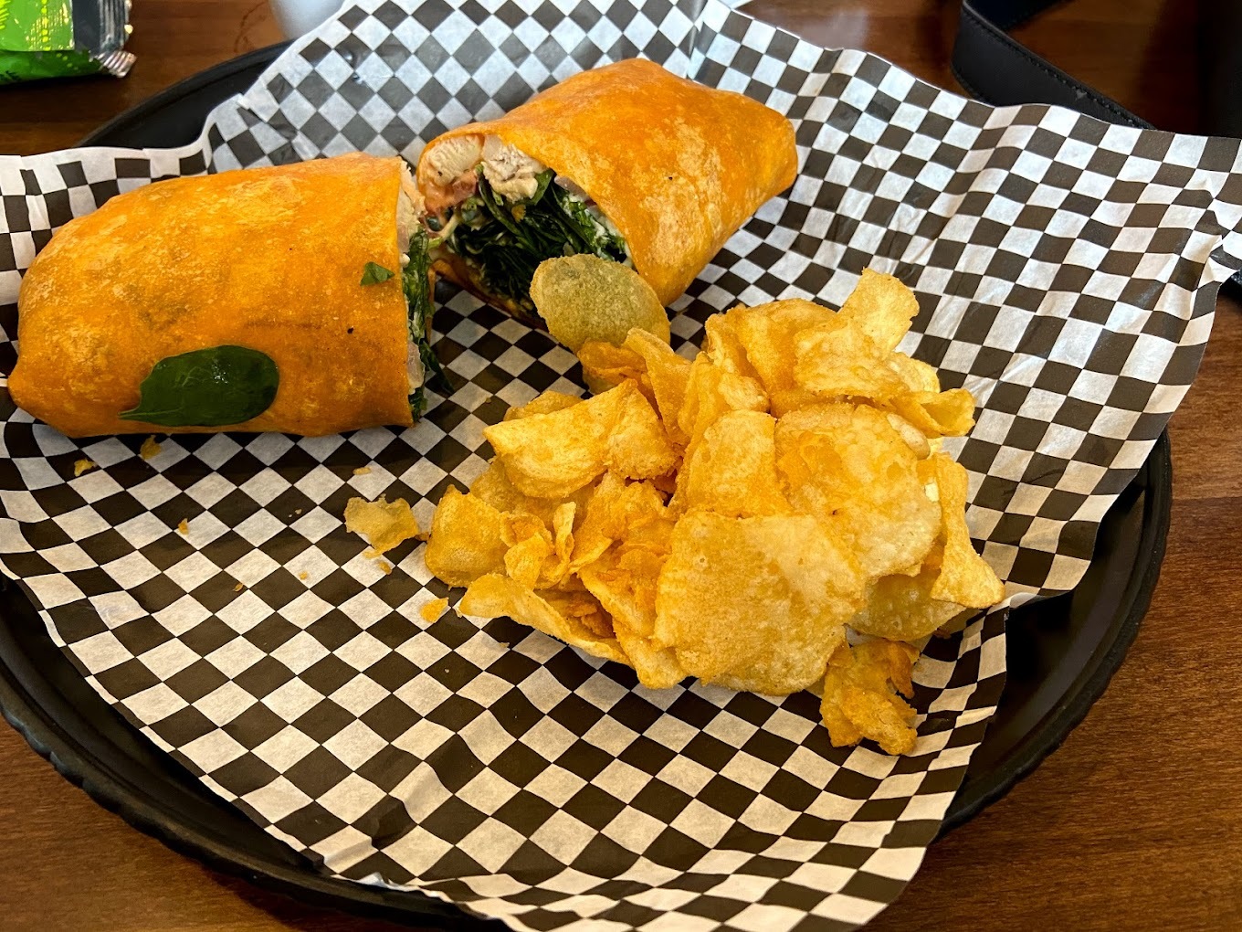 A basket with black and white checkered paper consisting of a sandwich with meat, spinach and sauce with a side of kettle chips.