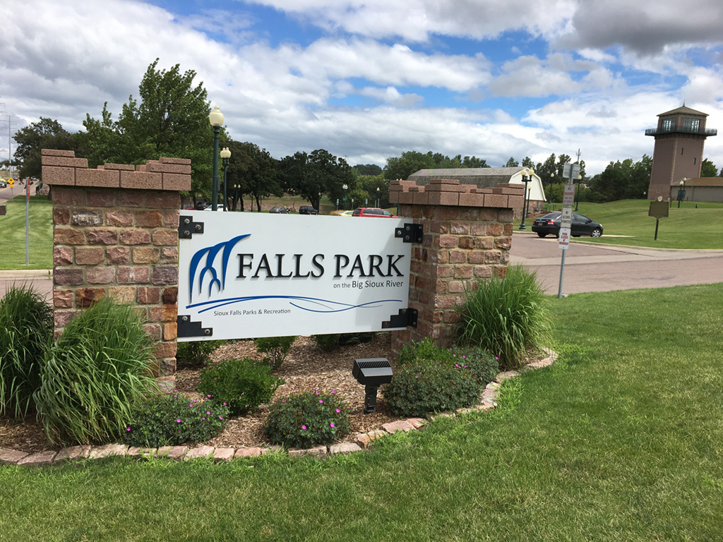 Falls park sign with two short brick columns and greenery bushes.