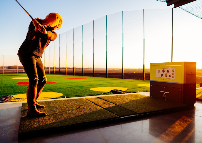 Woman swinging a golf club, about to hit a golf ball into the golfing arena during sunset.