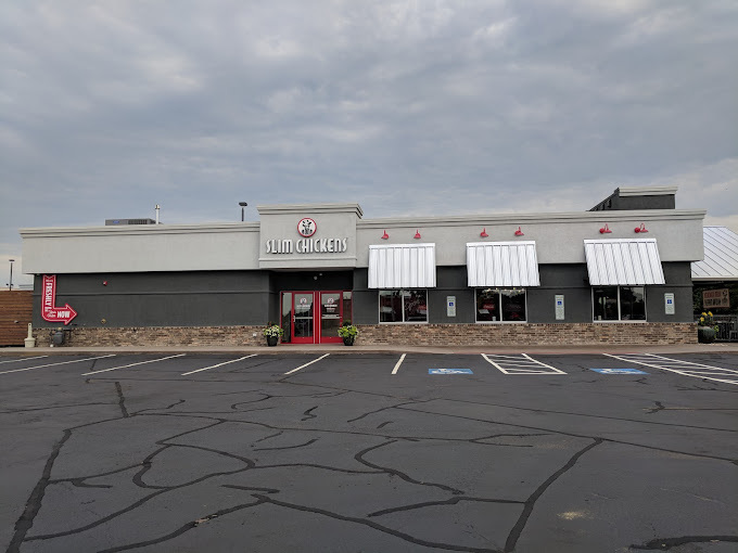 Faraway exterior view of a grey and beige building with white sunshades over the windows. The entrance doors are red and have a sign above it that says "Slim Chickens."