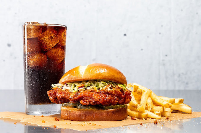 Burger with a large chicken patty and shredded green, a stack of french fries, and a glass of a dark-colored drink with ice. This sets on a piece of parchment paper with quite a few food crumbs.