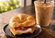 Flaky croissant breakfast sandwich with bacon, egg, and melted cheese served beside a creamy iced coffee on a wooden café table.