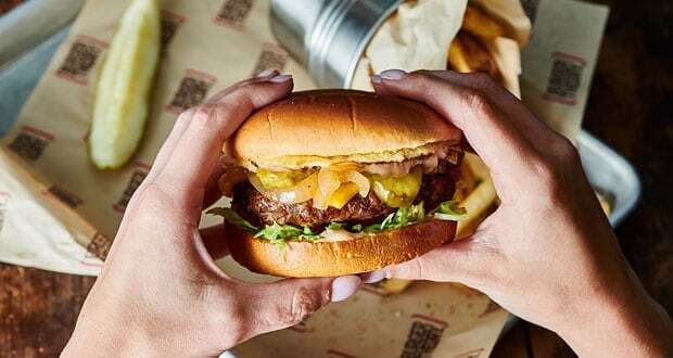 A meal tray with a pickle and fries in the background of a man holding a large burger with both hands.