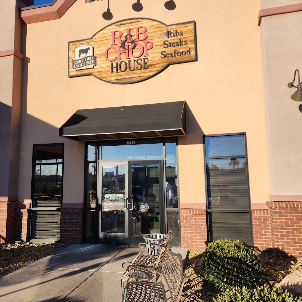 The entrance to a restaurant with beige colored siding, a faux wooden sign is above the black doors reading "Rib & Chop House" in red and black lettering.
