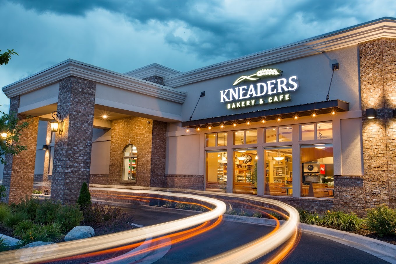 A tan building with an drive up awning, above the awning on the building is a large lighted sign reading "Kneaders"