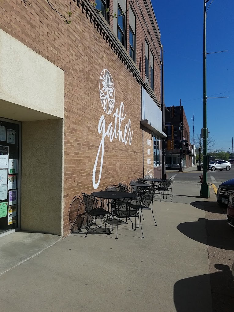 An outside view of a cafe with brick walls and a large sign with the lettering Gather in white cursive. Outdoor seating is also shown.
