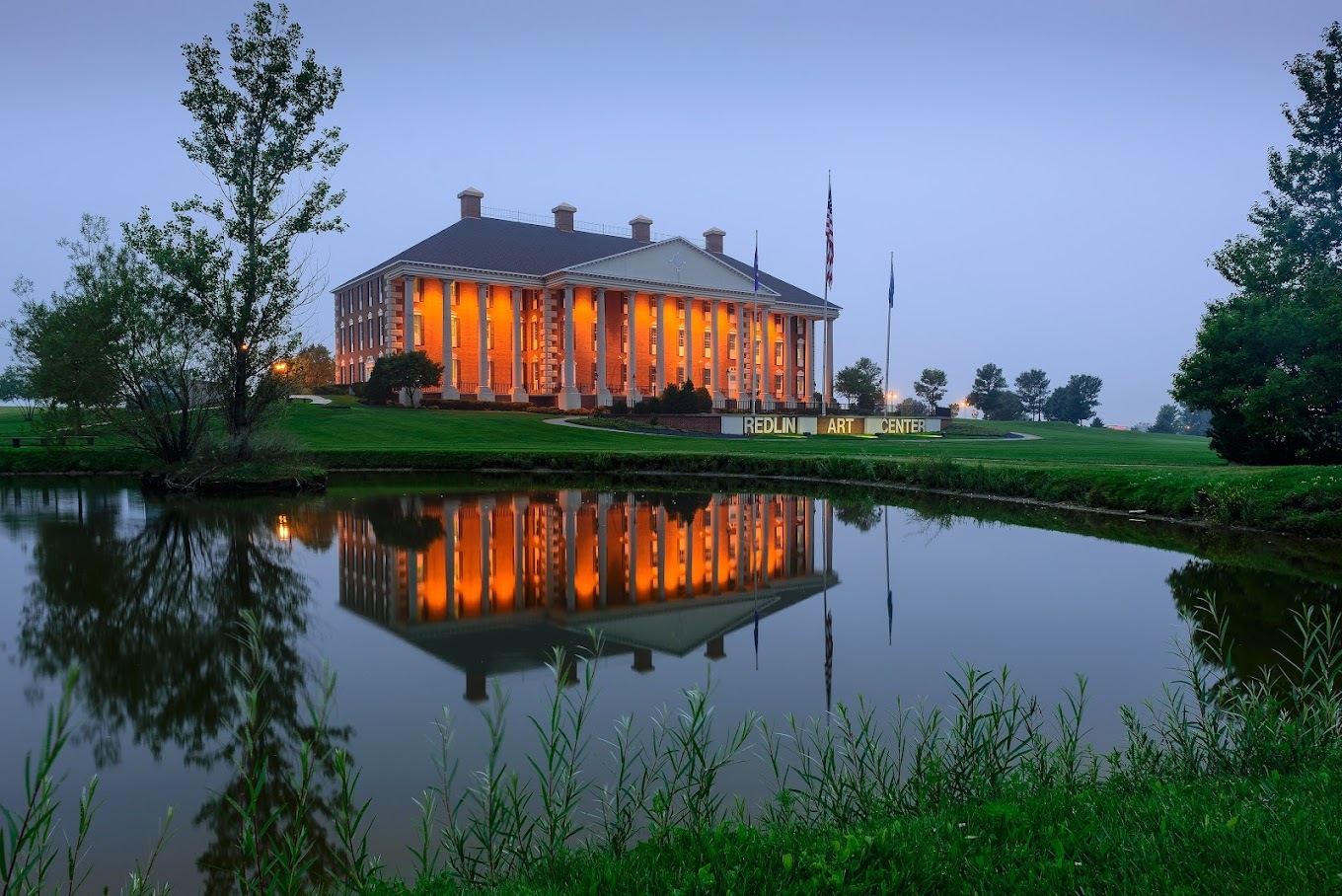 A landscape photo of a pond in front of a large brick estate with an awning and large white pillars.