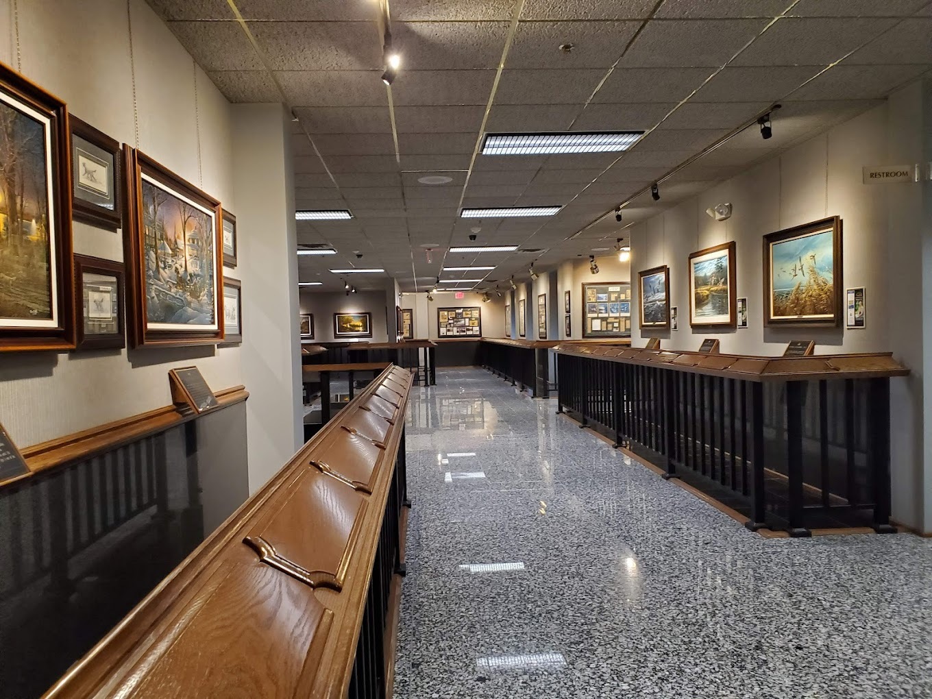 A long hallway with many paintings lighted, with wooden barriers in front of the paintings.